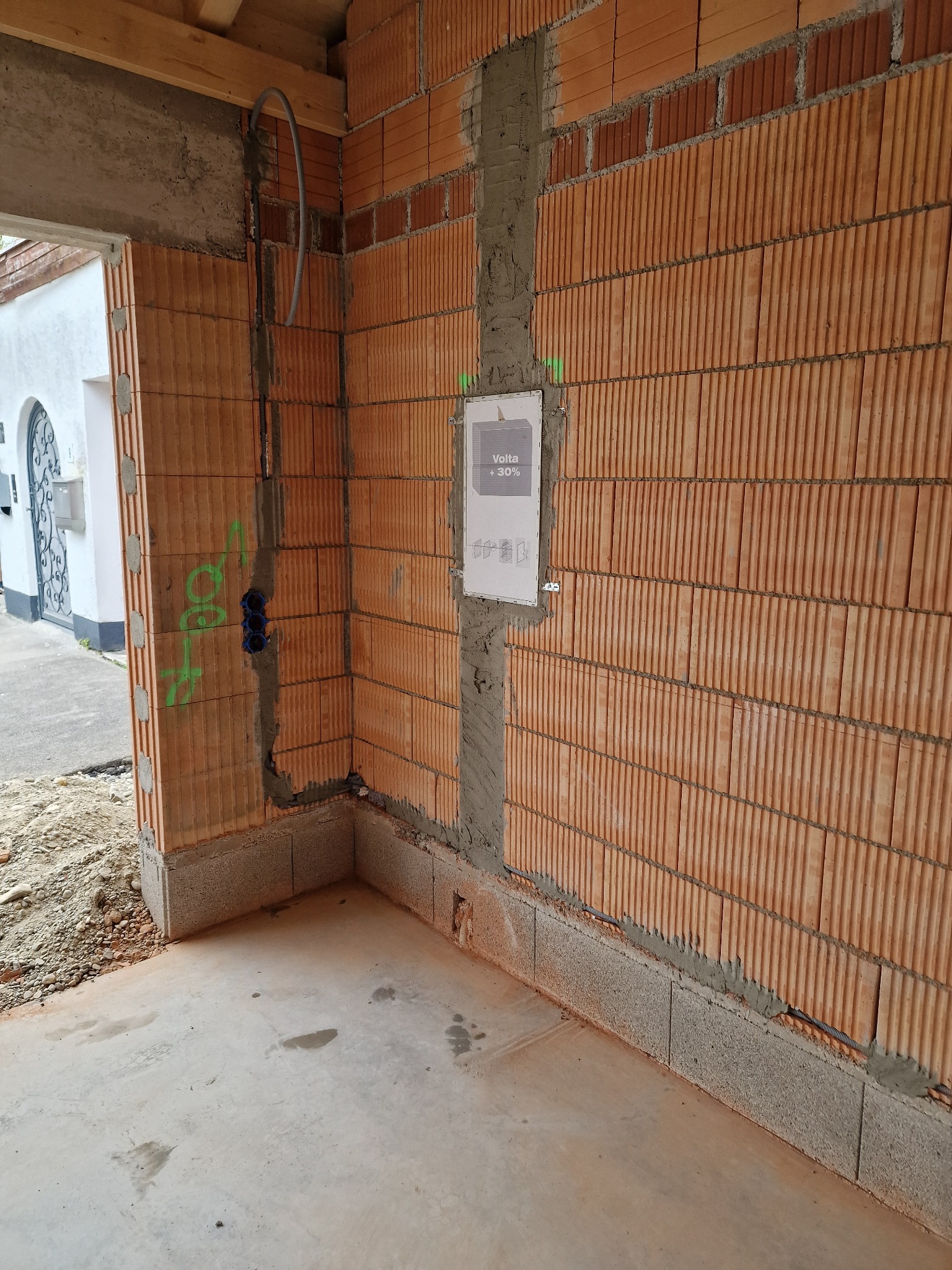 Surowy stan wnętrza budynku w trakcie prac murarskich, widoczne pomarańczowe cegły dziurawki, szare spoiny, pionowy peszel z przewodami i puszki instalacyjne w ścianie, oznaczenia zielonym sprayem.