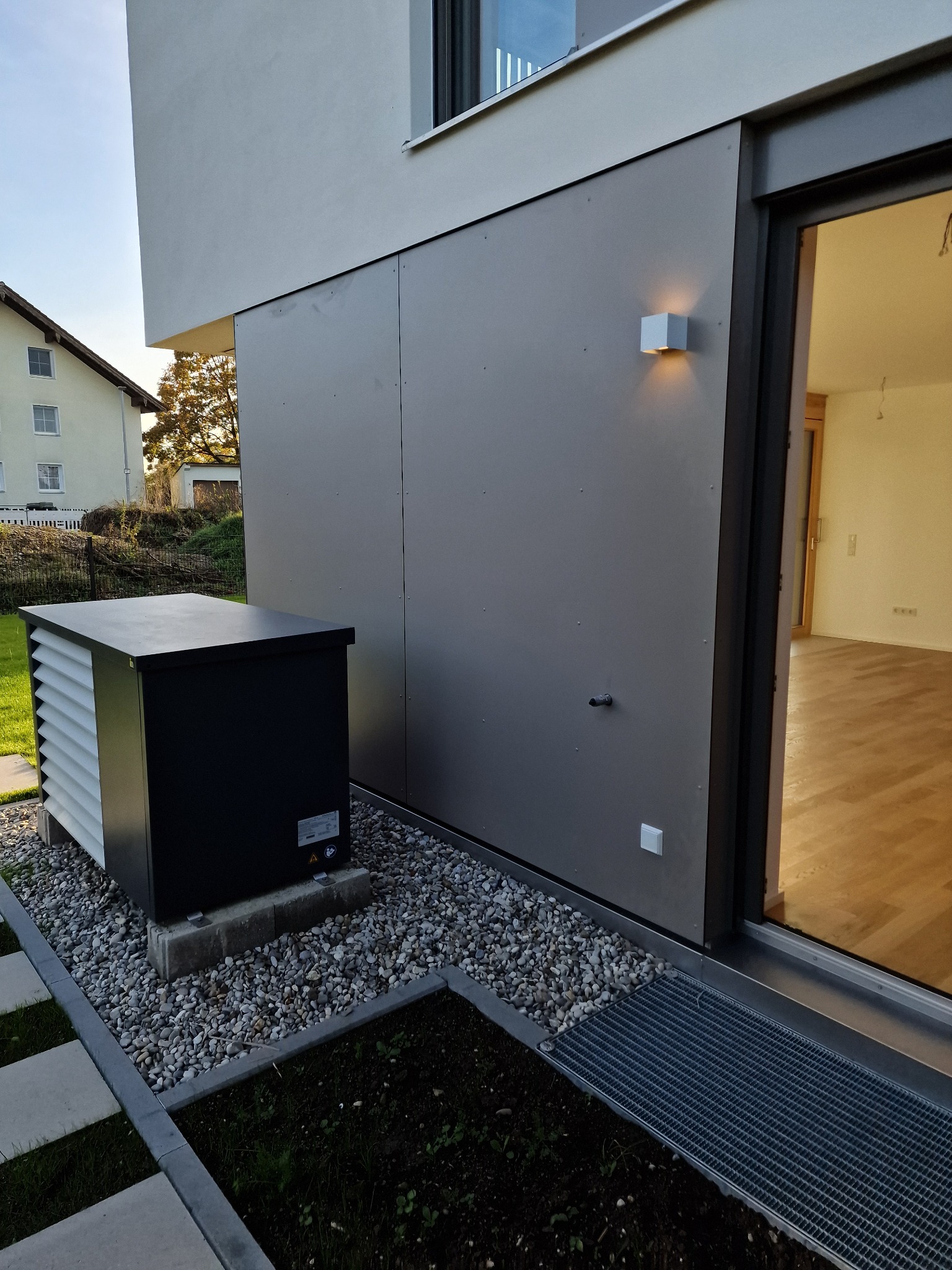 Zewnętrzna jednostka pompy ciepła, czarna obudowa z białymi żaluzjami, umieszczona na betonowych blokach na żwirowym podłożu, obok nowoczesnego budynku z szarą elewacją i kwadratowym oświetleniem.