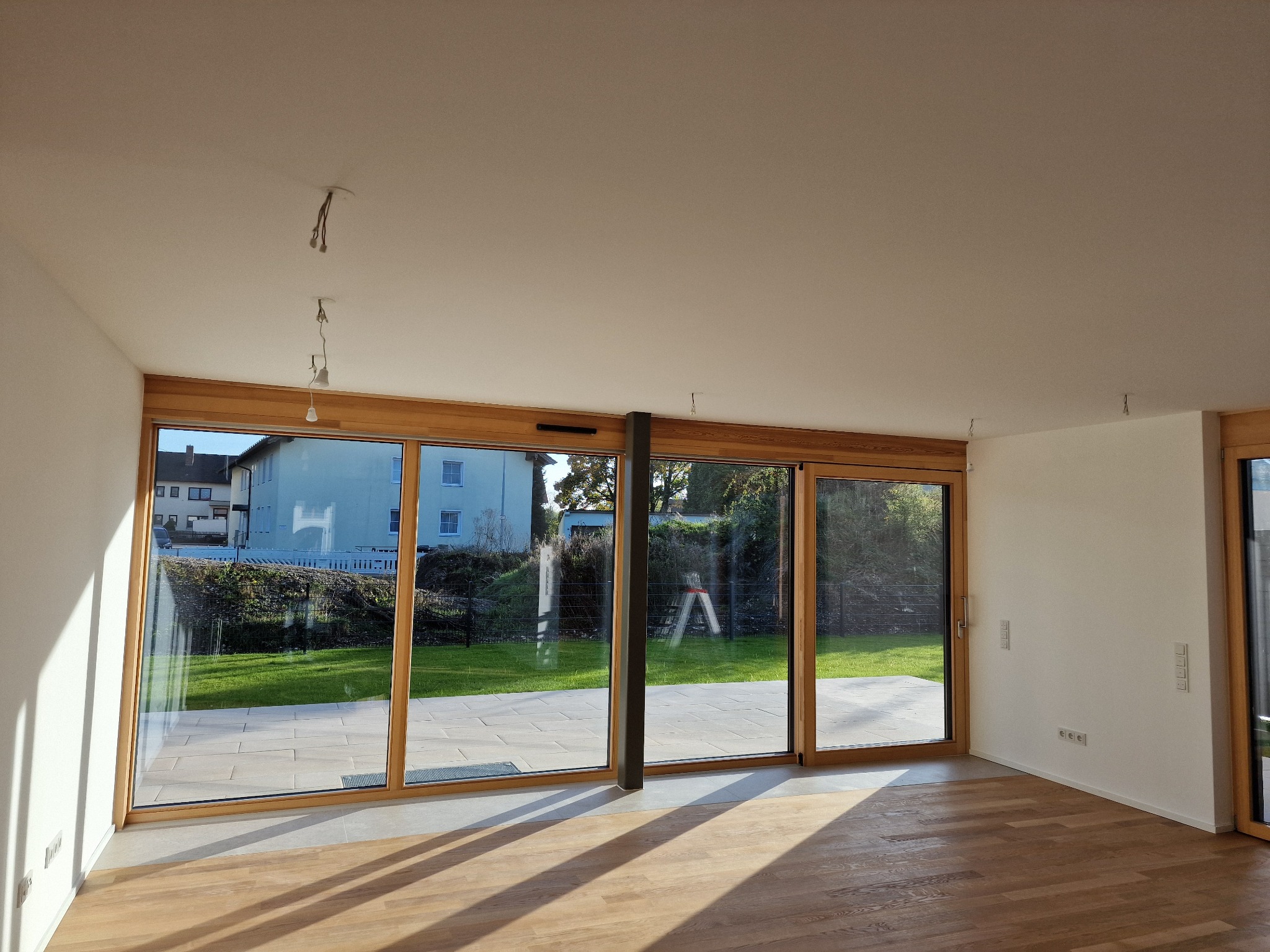 Jasne wnętrze z drewnianą podłogą i dużymi przesuwnymi oknami z widokiem na ogród; widoczne wyprowadzone przewody oświetleniowe w suficie.