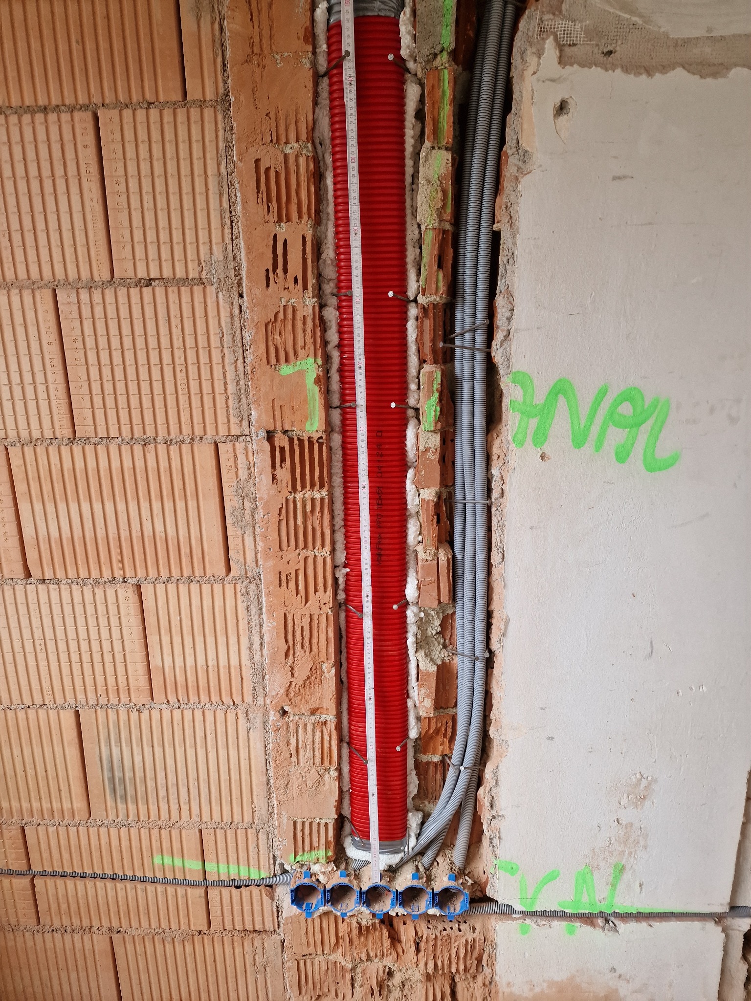 Wewnętrzne instalacje w trakcie budowy: czerwona rura karbowana pionowo zamocowana do ściany z cegły dziurawki z pomocą pianki montażowej, obok szare rury karbowane, na ścianie napis 'ANAL'...
