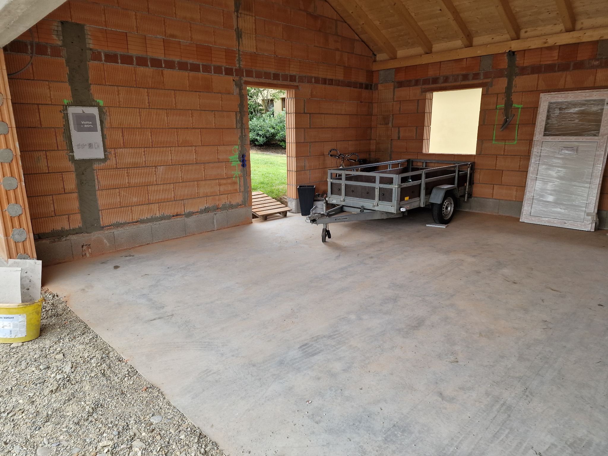 Wnętrze surowego garażu z czerwonej cegły z widocznymi puszkami elektrycznymi w ścianach, przyczepką samochodową i drzwiami foliowanymi, w tle widok na ogród.