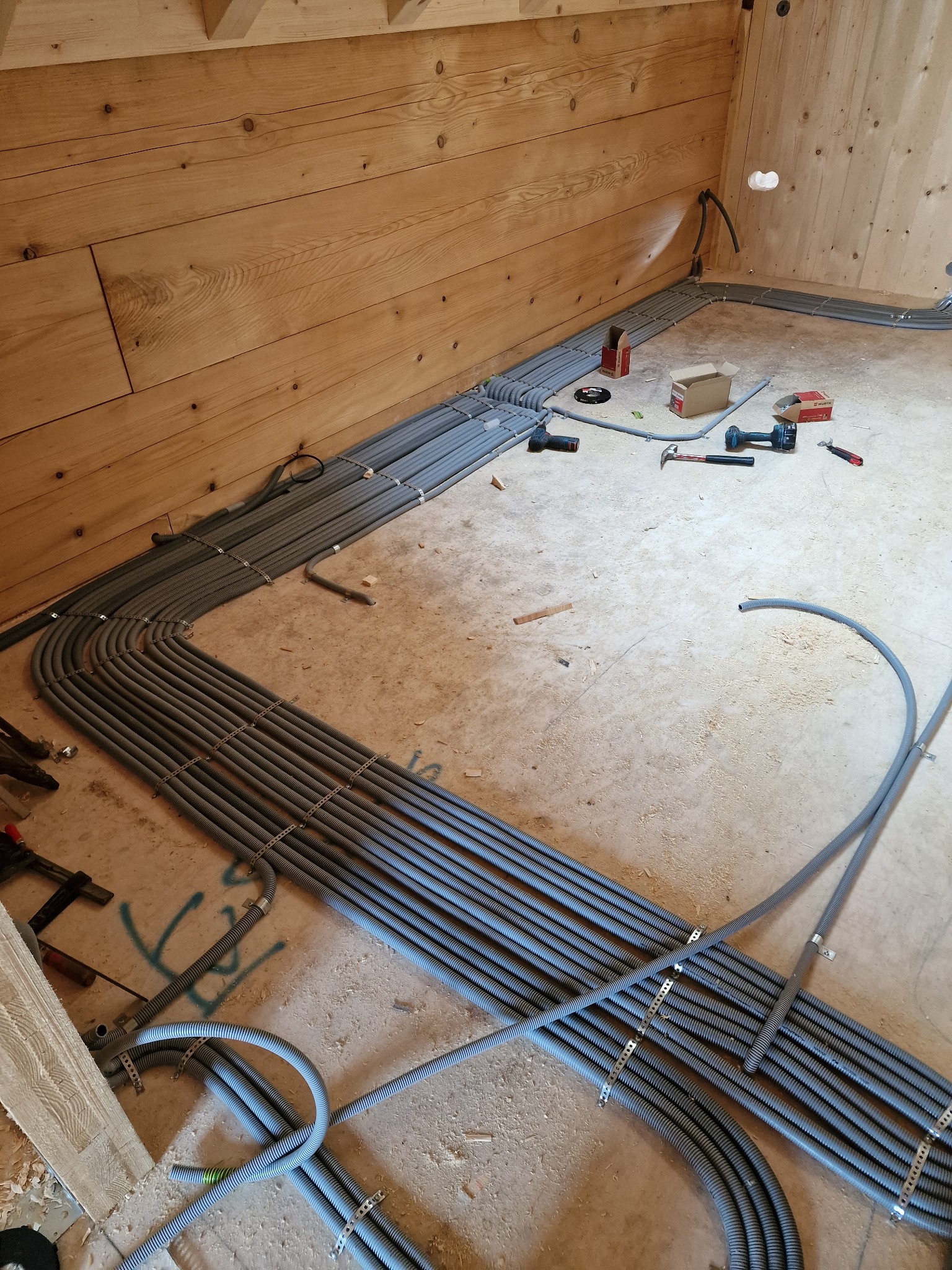 Szereg szarych, karbowanych rur ułożonych wzdłuż drewnianej ściany w surowym wnętrzu, zabezpieczonych metalowymi uchwytami, z widocznymi narzędziami i elementami montażowymi na podłodze.