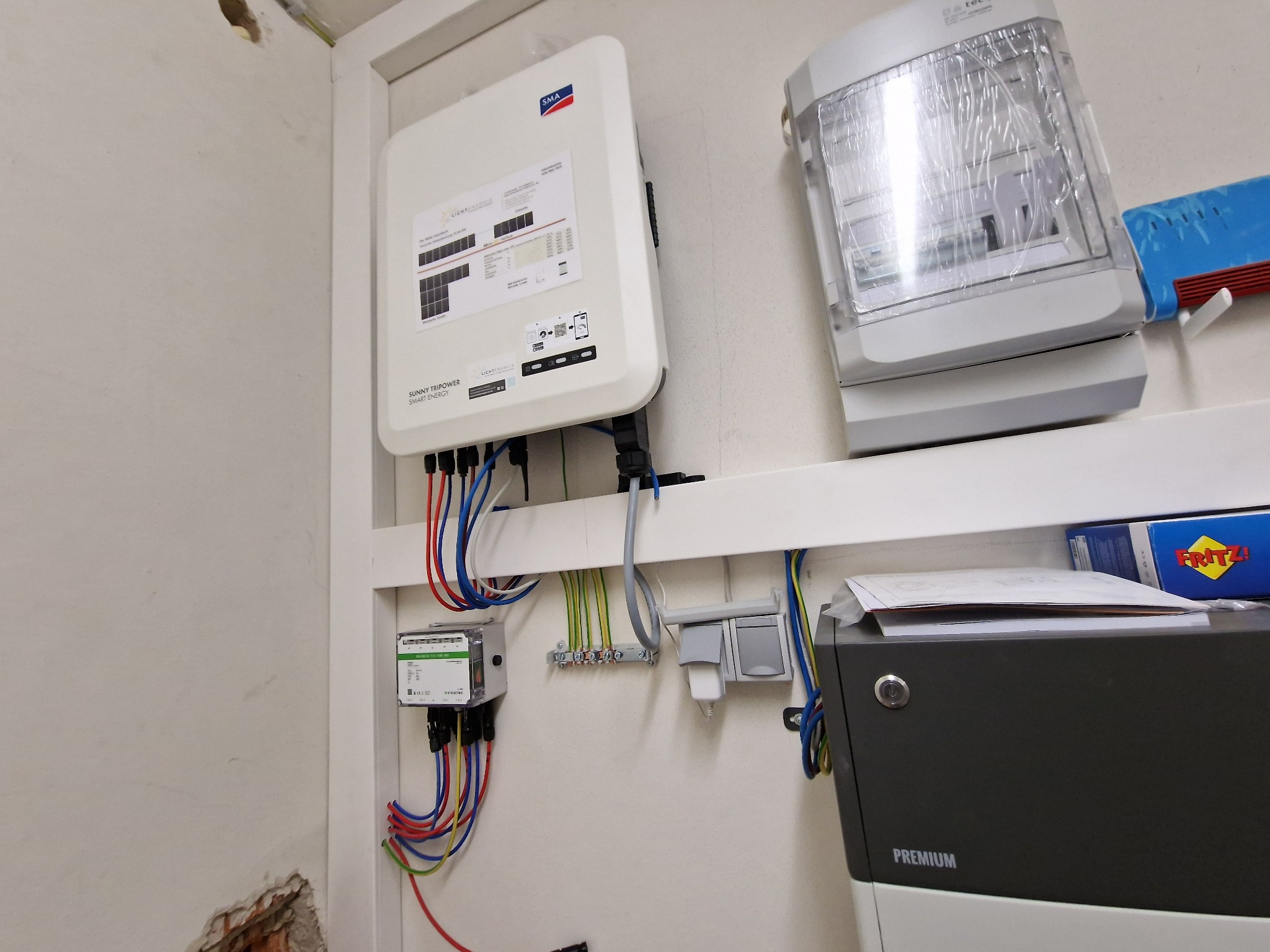 Biała skrzynka inwertera SMA zamontowana na ścianie obok licznika energii i zabezpieczonego plastikową folią oświetlenia, widoczne przewody i złącza, ściana z ubytkiem tynku w rogu.