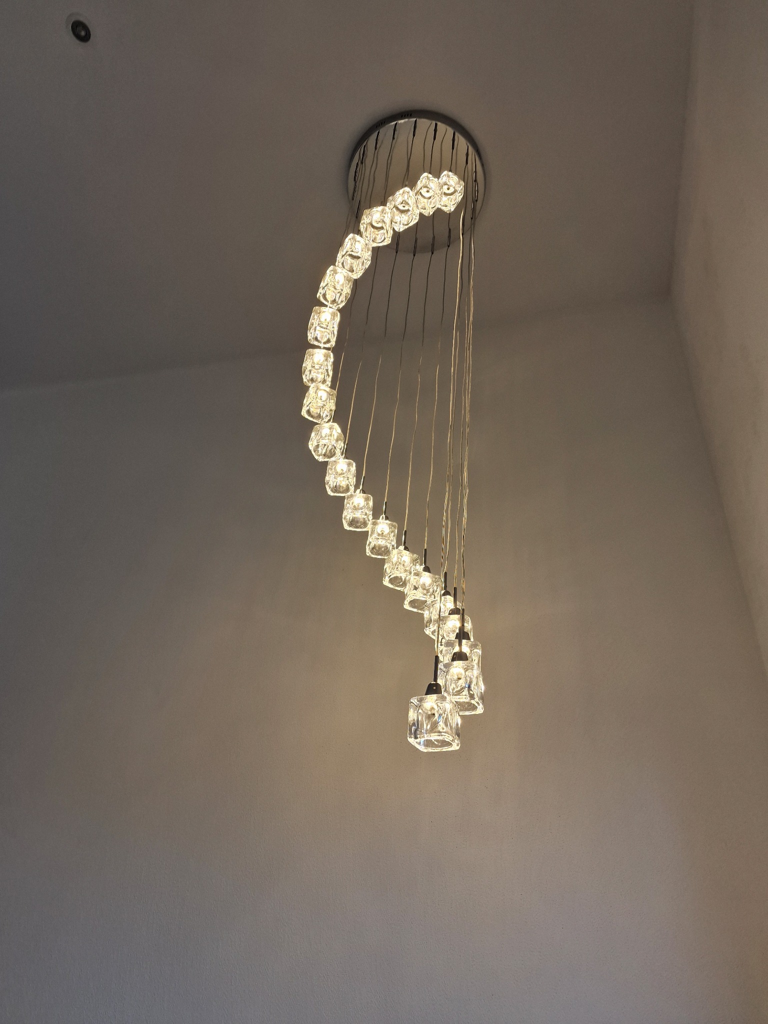 Spiralna lampa wisząca z kryształowymi elementami, zamontowana na suficie, oświetlona ciepłym światłem, widok od dołu.