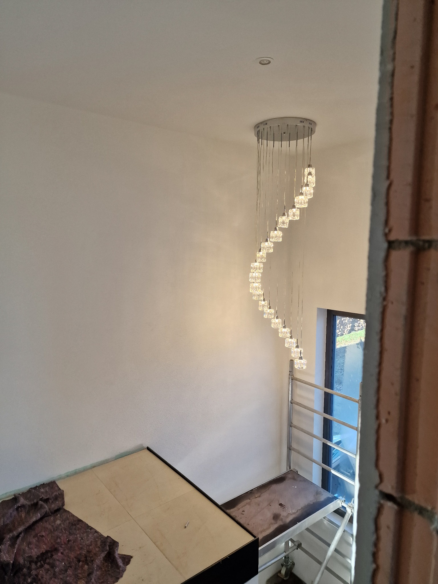 Spiralny żyrandol z kryształkami, widoczny z dołu, zamontowany w nowym domu w trakcie wykańczania, z rusztowaniem i zabezpieczeniem podłogi.
