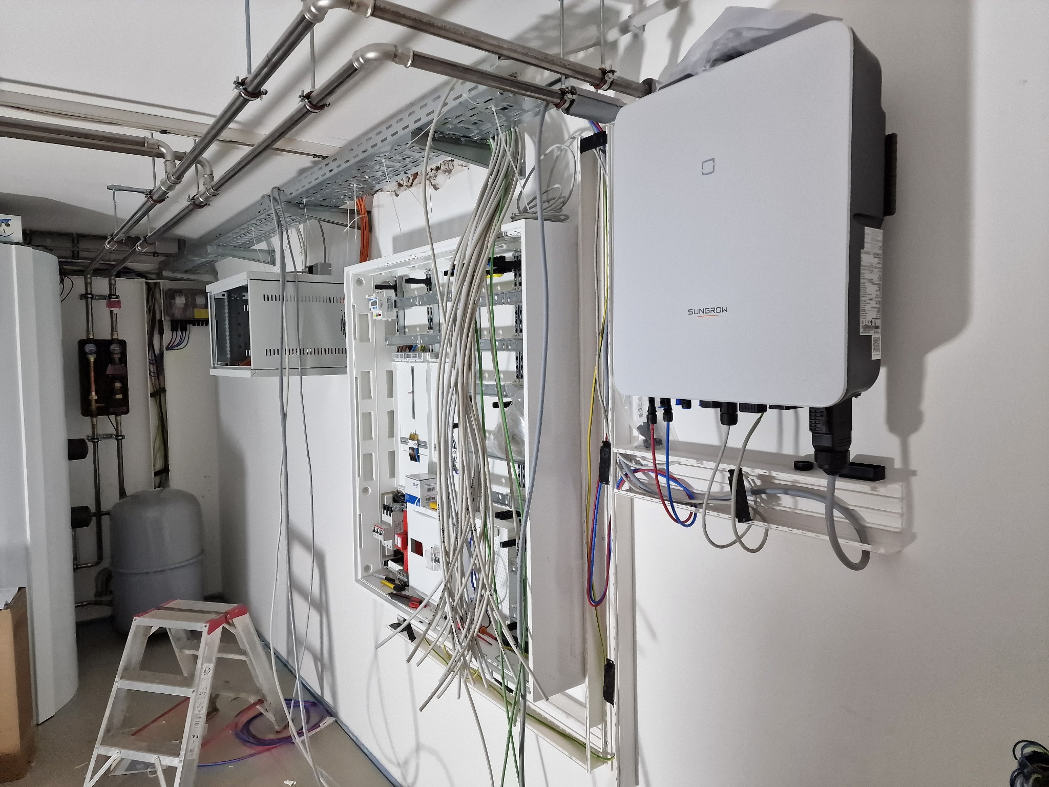 Wnętrze kotłowni w trakcie montażu instalacji elektrycznej: biała skrzynka rozdzielcza z przewodami, szara drabina, biały inwerter Sungrow na ścianie, rury instalacyjne pod sufitem.
