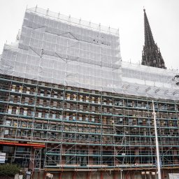 JTM-BAU SPÓŁKA Z OGRANICZONĄ ODPOWIEDZIALNOŚCIĄ - Budynek w rusztowaniach, zabezpieczony siatką ochronną, w tle wieża kościoła i nowoczesny budynek biurowy. Widoczny dźwig budowlany.