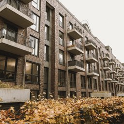 JTM-BAU SPÓŁKA Z OGRANICZONĄ ODPOWIEDZIALNOŚCIĄ - Nowoczesny budynek wielorodzinny z ceglaną elewacją, balkonami z metalowymi balustradami i donicami z roślinami wzdłuż ogrodzenia.