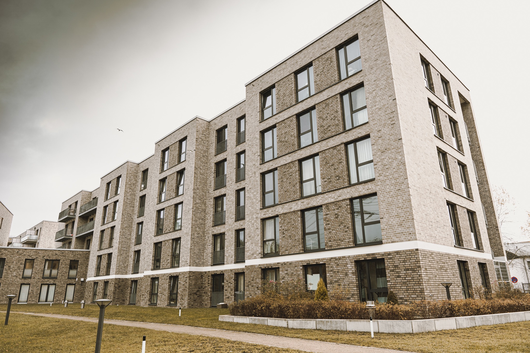 Nowoczesny budynek mieszkalny z elewacją z jasnej cegły, widok z dołu na pochmurnym niebie, z widocznymi balkonami i elementami zieleni.