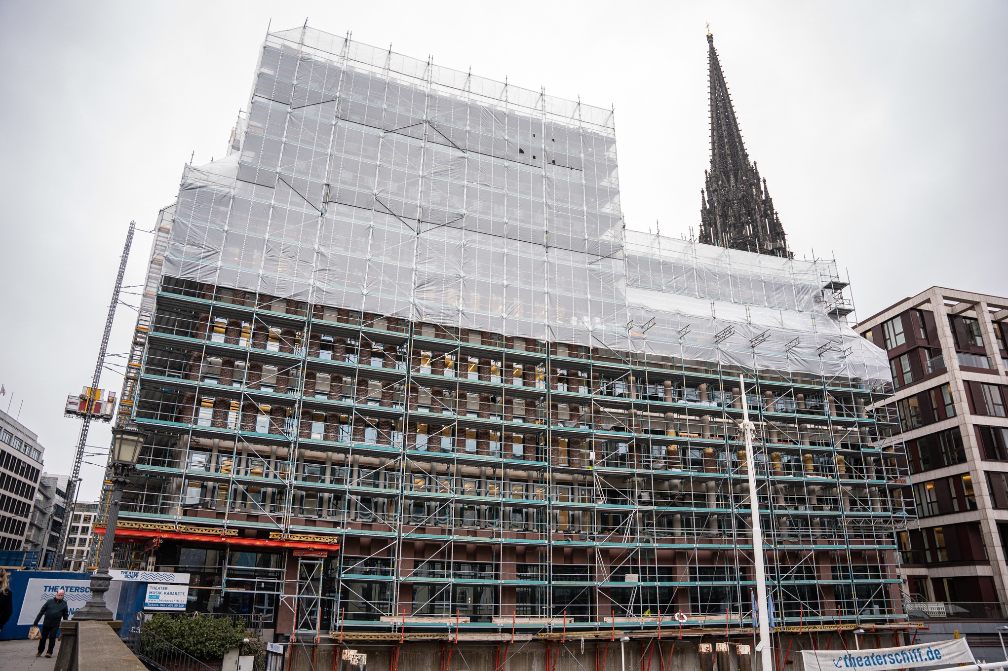Budynek w rusztowaniach, zabezpieczony siatką ochronną, w tle wieża kościoła i nowoczesny budynek biurowy. Widoczny dźwig budowlany.