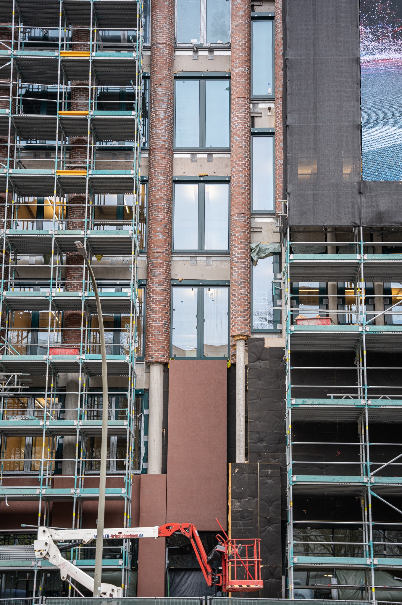 Budynek w trakcie prac elewacyjnych: widoczne rusztowania, fragmenty elewacji z cegły, okna, oraz obszary przygotowane do ocieplenia, z użyciem podnośnika koszowego.