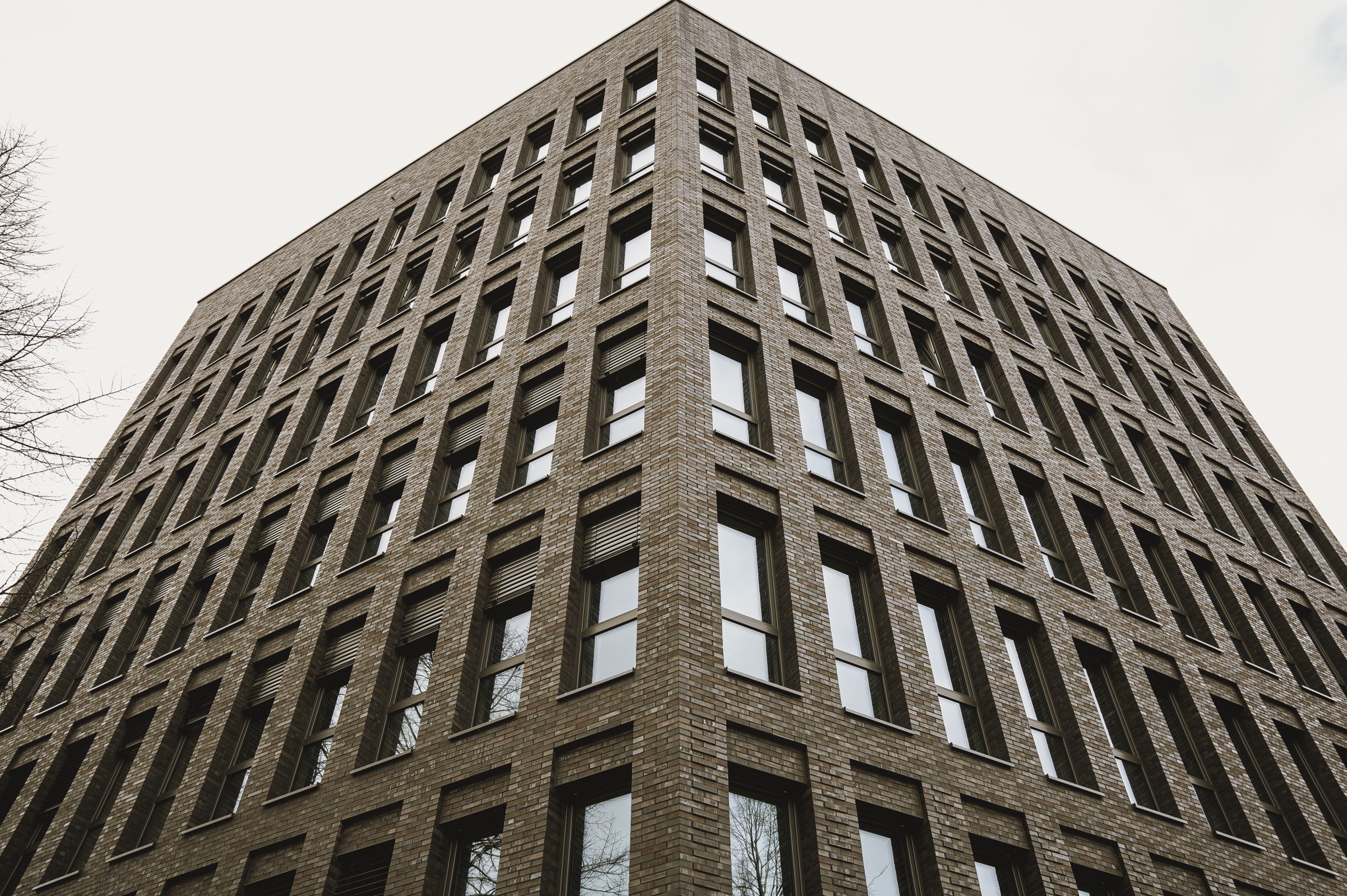 Wielopiętrowy budynek z ceglaną elewacją, widok z dołu ukazujący liczne okna z ciemnymi ramami i częściowo zasłoniętymi żaluzjami, na tle pochmurnego nieba.