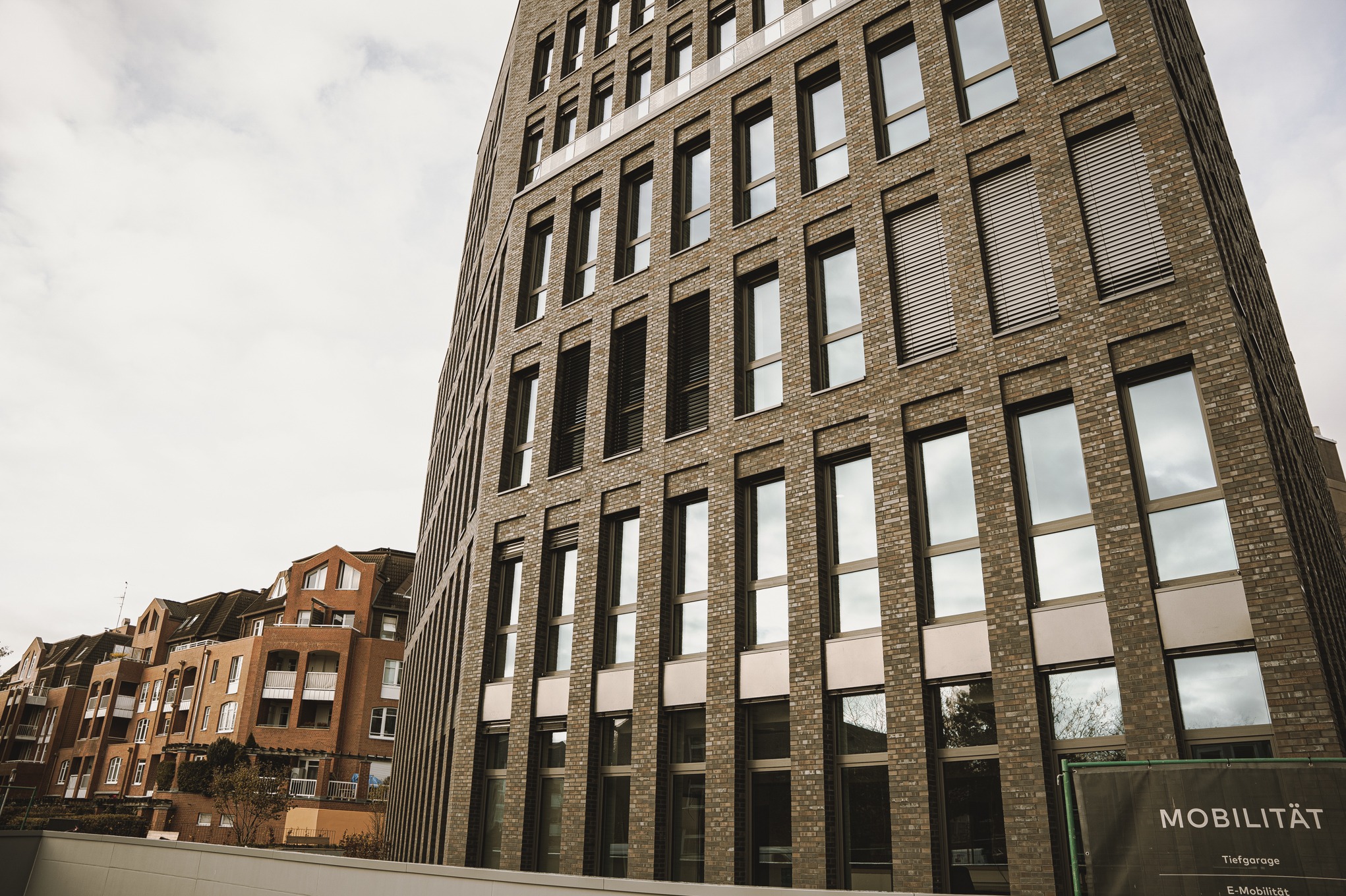 Nowoczesny budynek biurowy z ceglaną elewacją i dużymi oknami, widok z dołu na tle pochmurnego nieba, z widocznymi fragmentami sąsiedniej zabudowy.