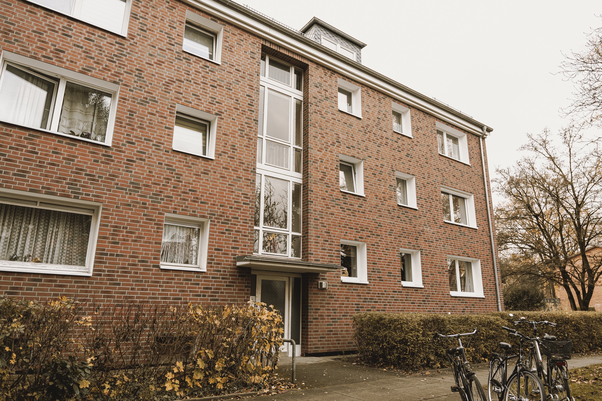 Trzypiętrowy budynek mieszkalny z czerwonej cegły z białymi ramami okiennymi, widok z dołu na elewację, z rowerami zaparkowanymi przed budynkiem i krzewami w jesiennych barwach.