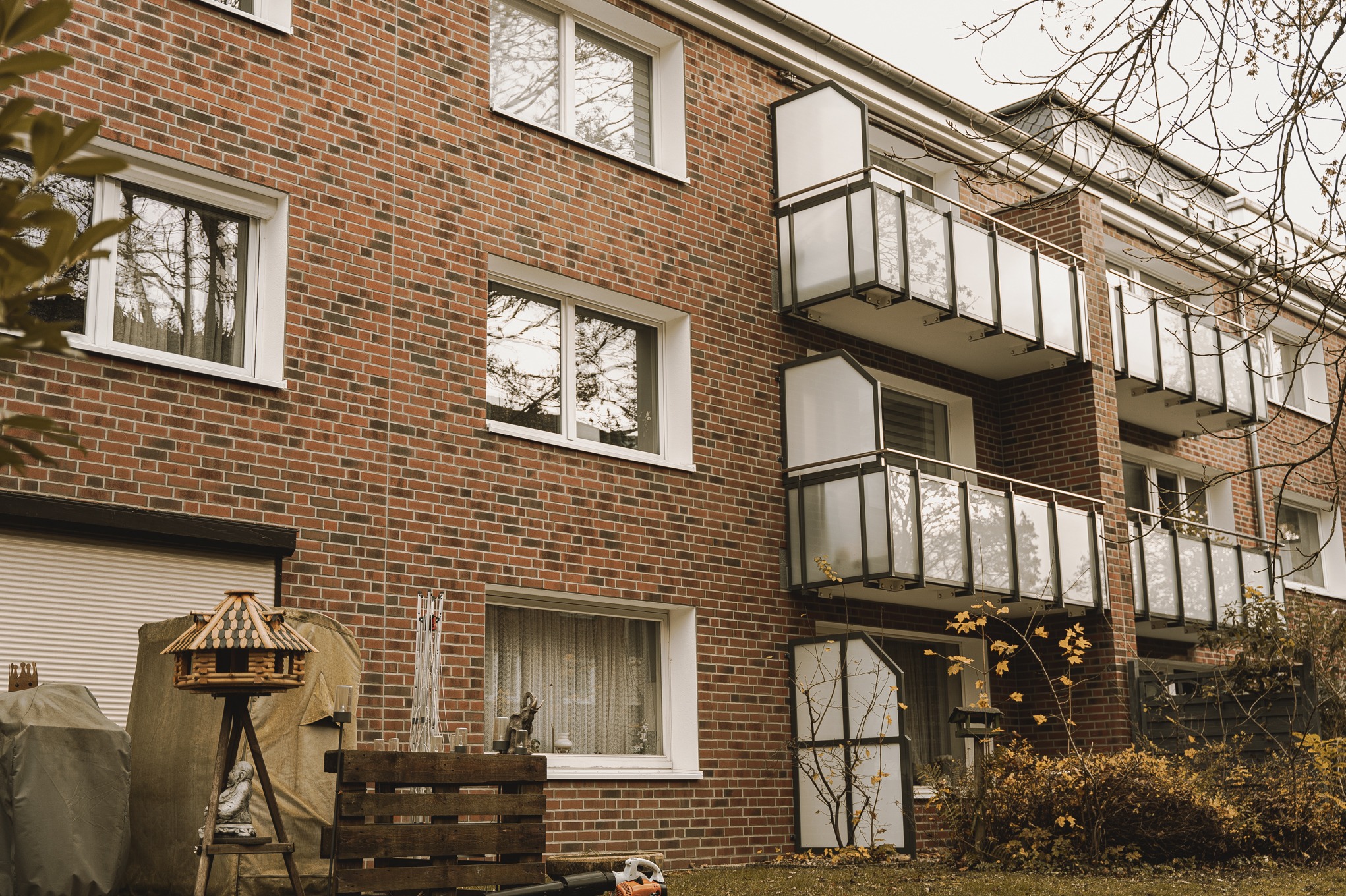 Czerwona ceglana elewacja budynku mieszkalnego z balkonami z białymi panelami, widok na parter z elementami dekoracyjnymi na trawniku.