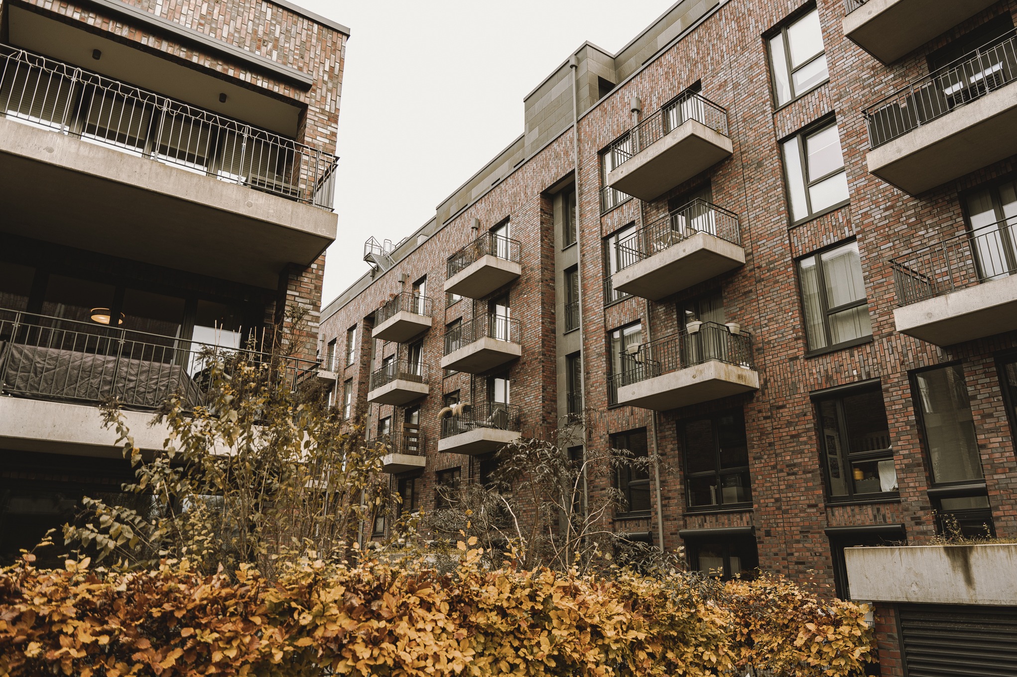 Nowoczesny budynek wielorodzinny z ceglaną elewacją, balkonami z metalowymi balustradami i widoczną roślinnością na pierwszym planie.