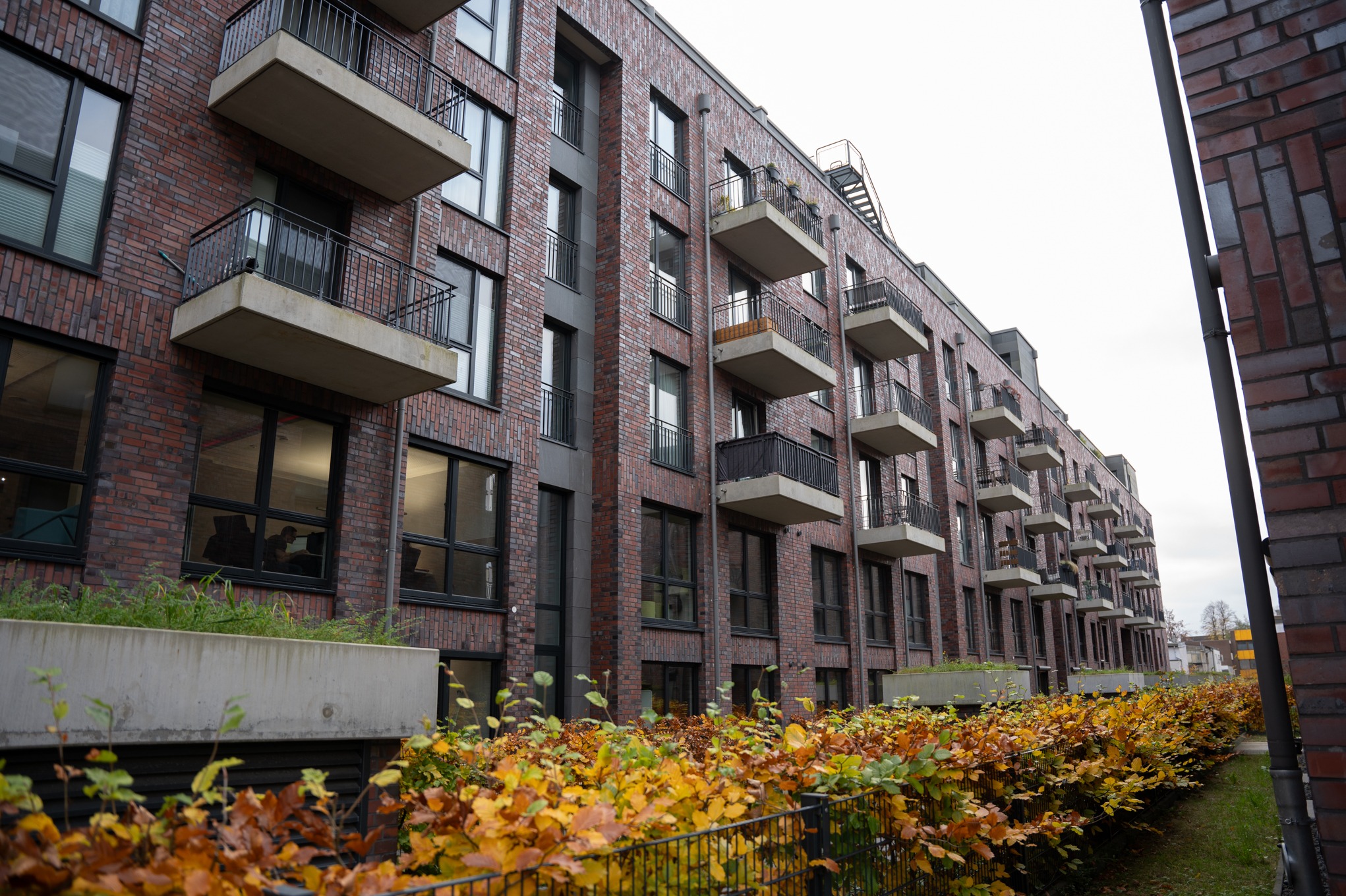 Nowoczesny budynek wielorodzinny z elewacją z czerwonej cegły, balkonami z szarą balustradą i betonowymi płytami, widok z poziomu gruntu na tle jesiennej roślinności.