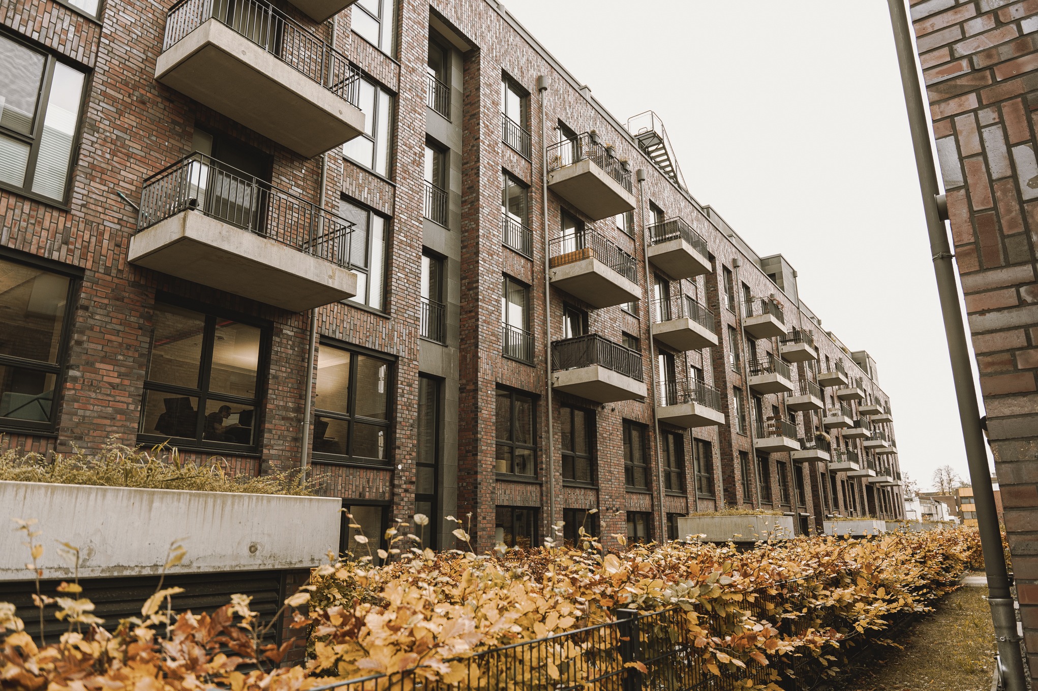 Nowoczesny budynek wielorodzinny z ceglaną elewacją, balkonami z metalowymi balustradami i donicami z roślinami wzdłuż ogrodzenia.