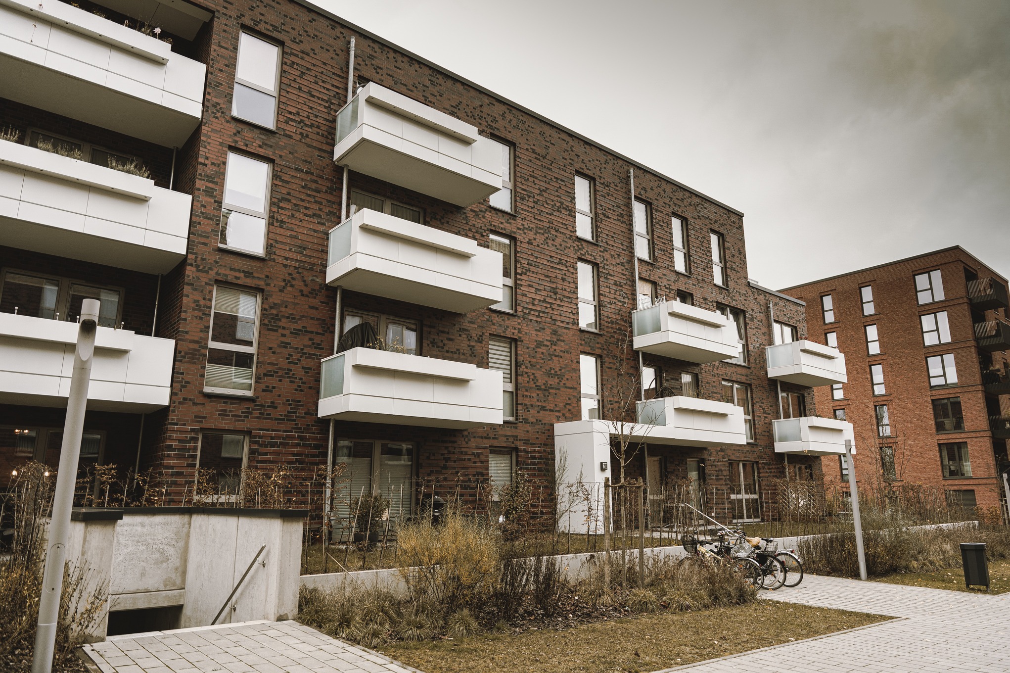 Nowoczesny budynek wielorodzinny z ceglaną elewacją, białymi balkonami i widocznymi rowerami przed budynkiem, w pochmurny dzień.
