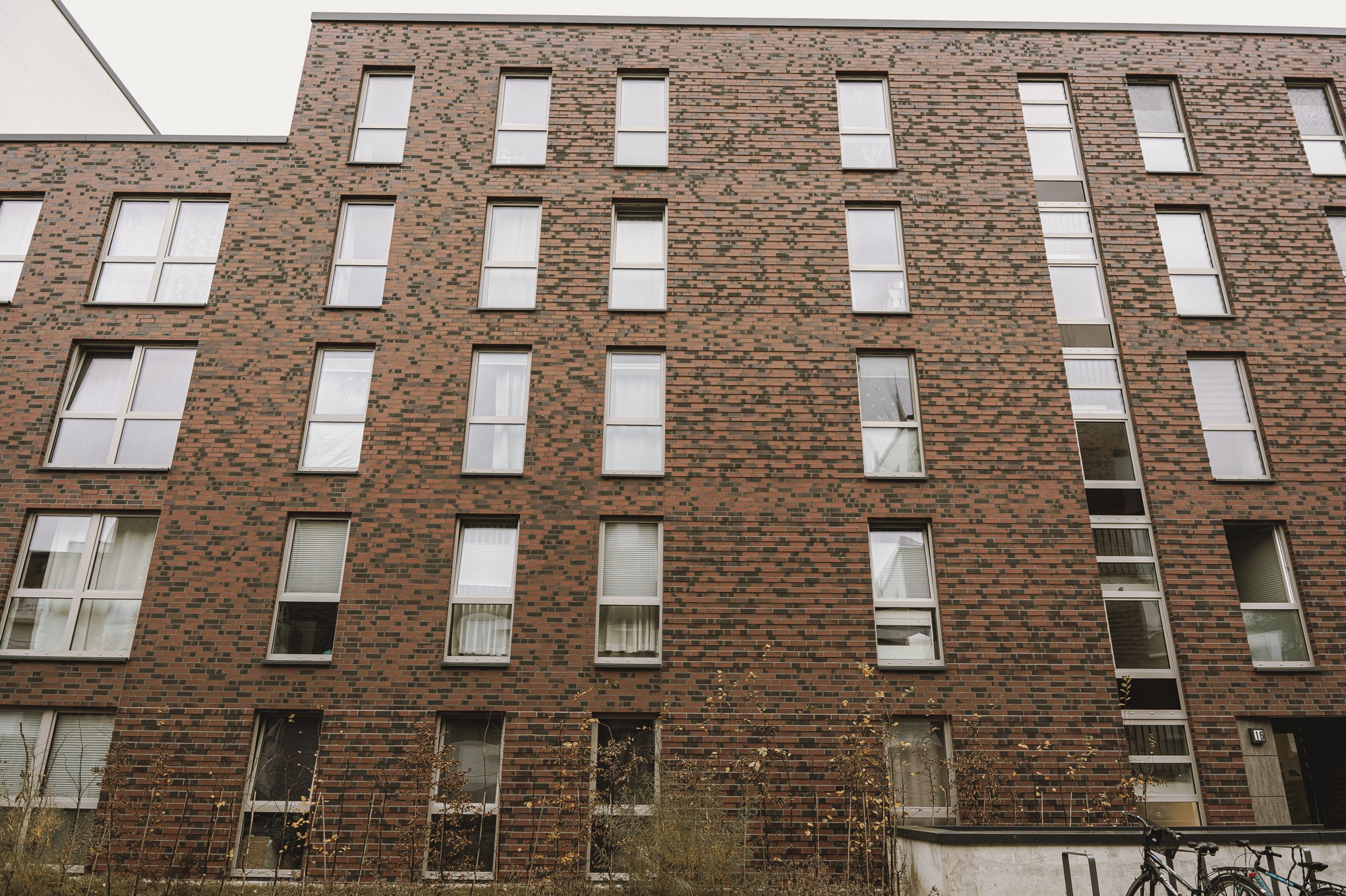 Wielopiętrowy budynek z elewacją z czerwonej cegły, widok z dołu, okna z białymi ramami, częściowo zasłonięte krzewami i rowerami.