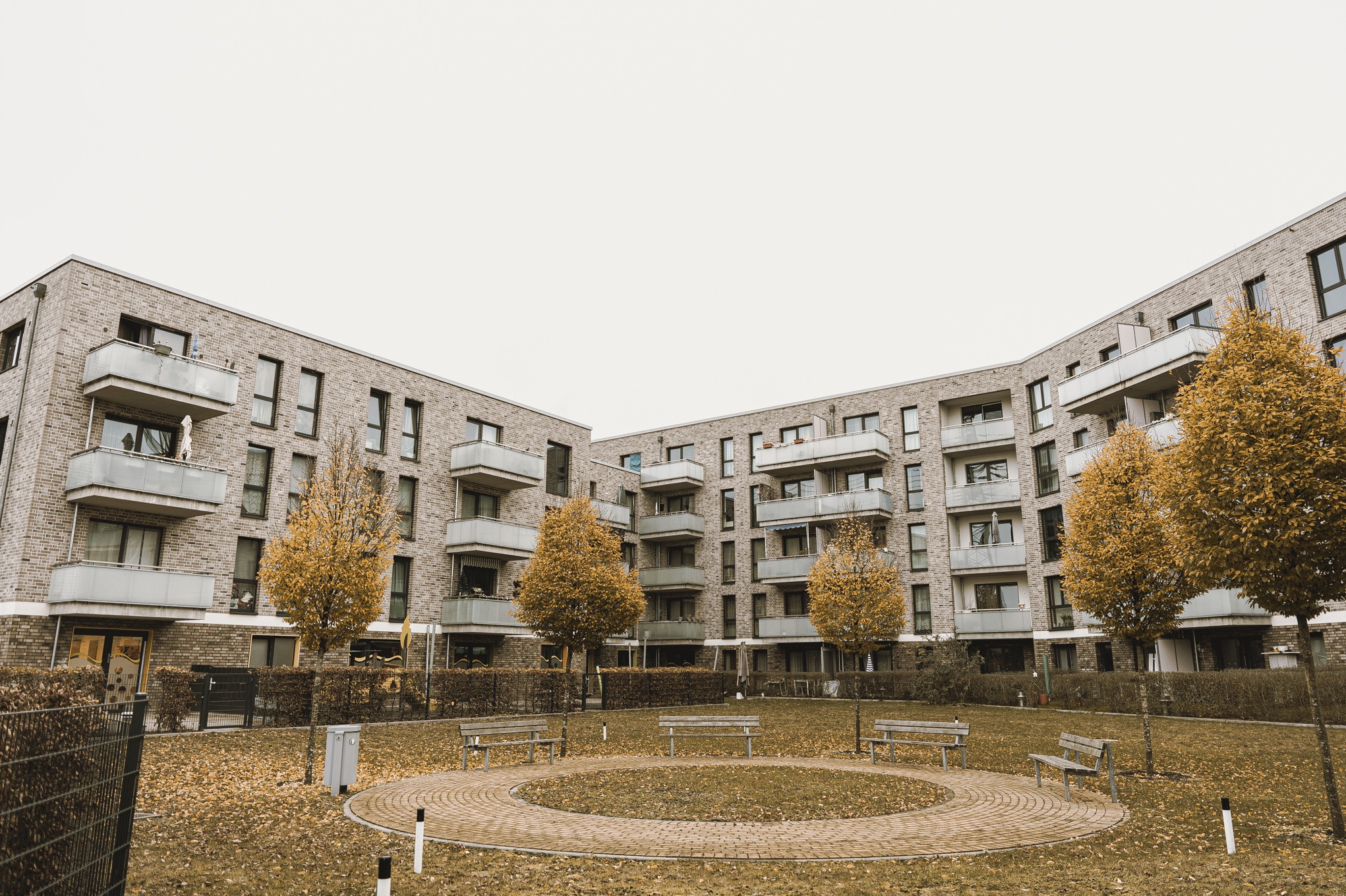 Nowoczesny budynek mieszkalny z jasnej cegły z balkonami, otoczony jesiennymi drzewami i parkiem z ławkami.