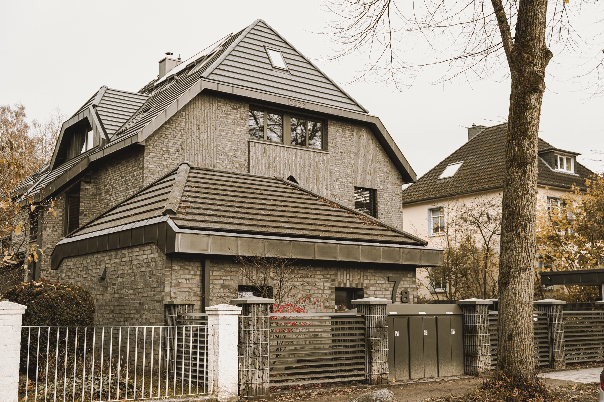 Dwupiętrowy dom z cegły klinkierowej z ciemną dachówką ceramiczną, widoczne ogrodzenie z metalowymi przęsłami i numer posesji 78.