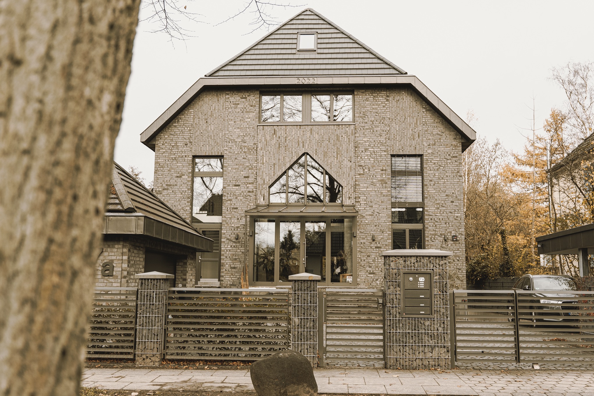 Nowoczesny dom z cegły klinkierowej, z dużymi oknami i oryginalnym wejściem, otoczony metalowym ogrodzeniem z elementami gabionowymi i drzewami jesienią w tle.