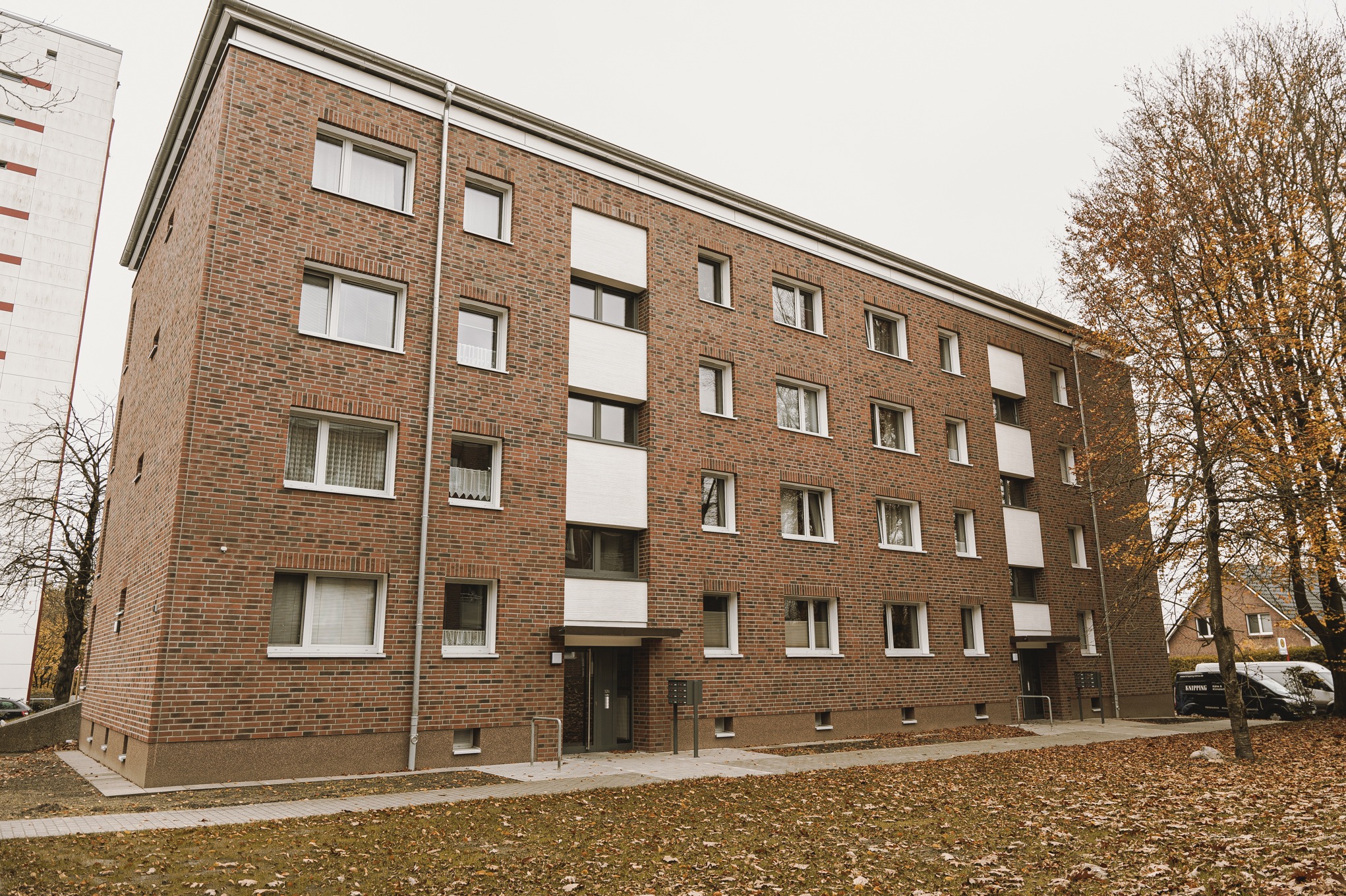 Czerwony ceglany budynek wielorodzinny z białymi ramami okiennymi i wejściami, widok z poziomu gruntu w jesiennej scenerii z opadłymi liśćmi.