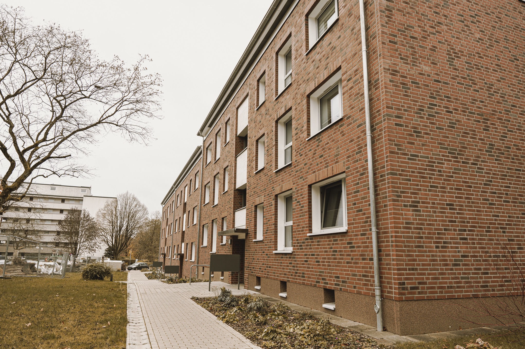 Długi budynek wielorodzinny z czerwonej cegły z białymi oknami, widok z poziomu chodnika, drzewo bez liści po lewej stronie.