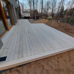 Surowy, jasny taras z desek z widocznym zarysem domu i placem budowy w tle.