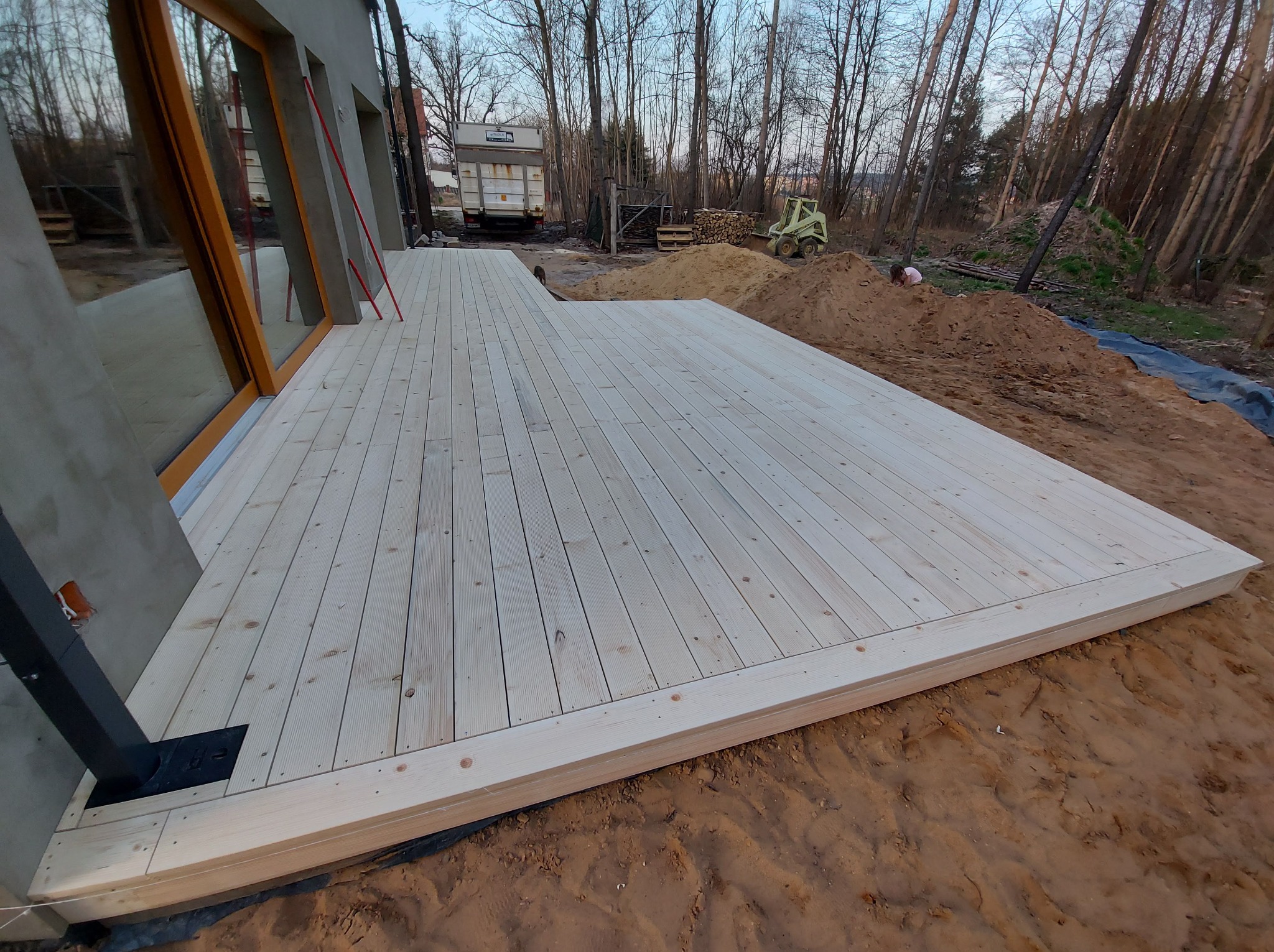 Surowy, jasny taras z desek z widocznym zarysem domu i placem budowy w tle.