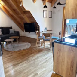 Jasne, drewniane podłogi w apartamencie z drewnianym sufitem, szarym narożnikiem, okrągłym dywanem, stołem z krzesłami i aneksem kuchennym.