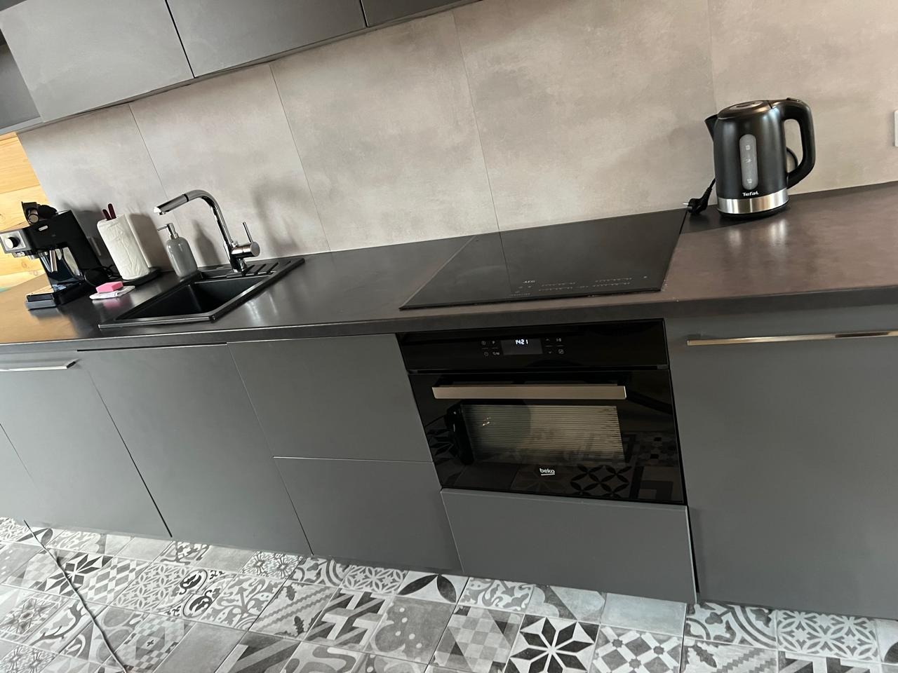 Nowoczesna szara kuchnia z błyszczącym blatem, czarnym zlewem i płytą indukcyjną, wzorzysta podłoga z płytek.