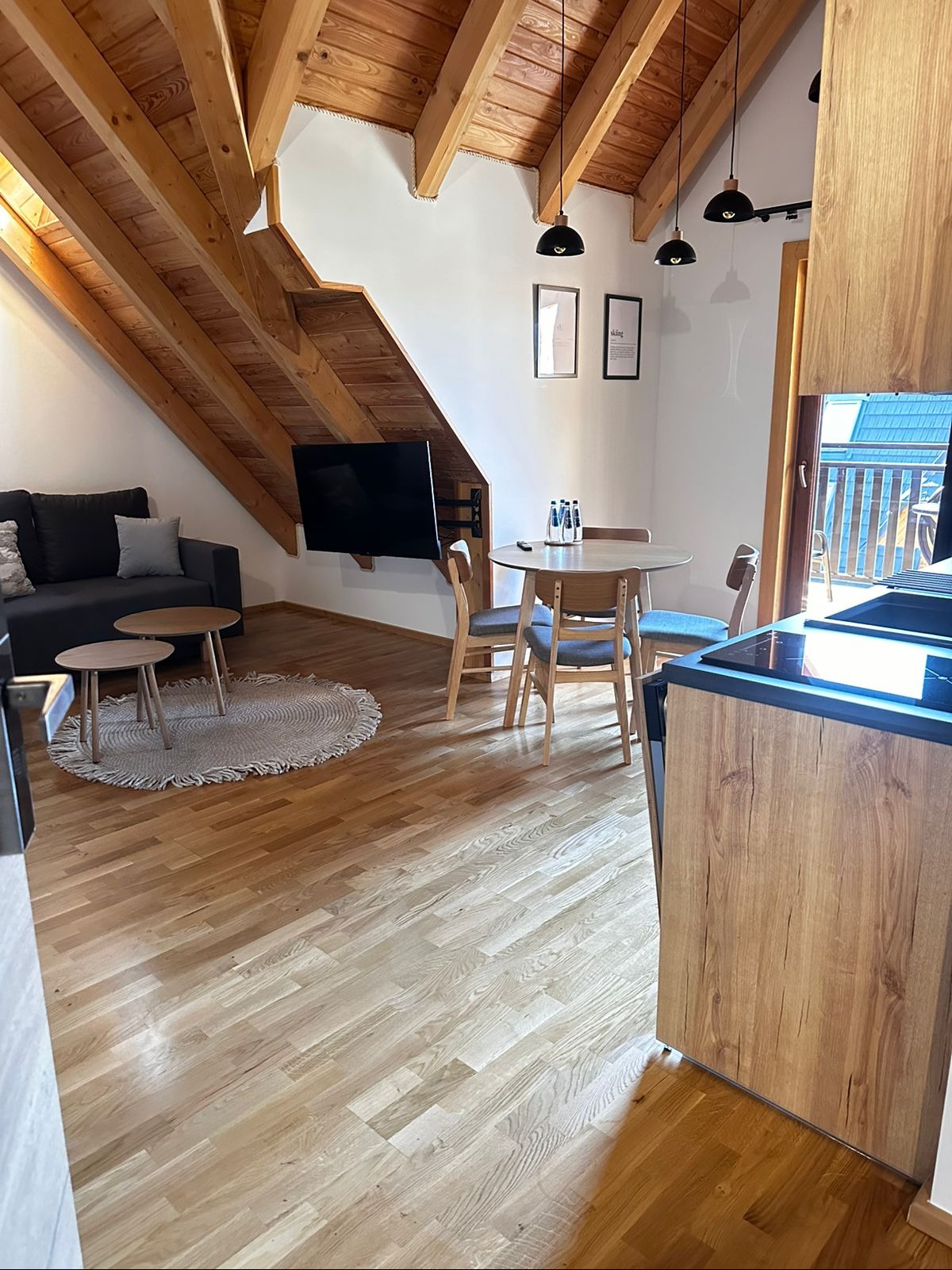 Jasne, drewniane podłogi w apartamencie z drewnianym sufitem, szarym narożnikiem, okrągłym dywanem, stołem z krzesłami i aneksem kuchennym.