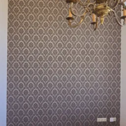 Ściana z tapetą w ornamenty w odcieniach szarości, parkiet na podłodze, fragment żyrandola i mebli. Widoczne gniazdka elektryczne przy podłodze.