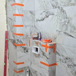TŁ Andrii Smaliuk - Łazienka w trakcie wykańczania: marmurowe płytki z pomarańczową taśmą, poziomice i narzędzia glazurnicze. Widoczny etap prac wykończeniowych.