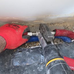 TŁ Andrii Smaliuk - Instalacja rur PEX z użyciem zaciskarki, widoczne rury w izolacji czerwonej i niebieskiej. Praca w trakcie wykonywania, rękawice ochronne.