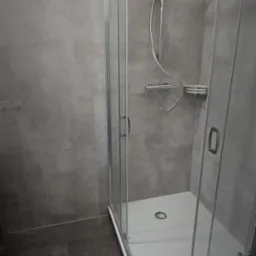 Nowoczesna kabina prysznicowa z szarymi płytkami imitującymi beton. Kabina narożna z chromowanymi detalami i ciemną podłogą. Minimalistyczny design.