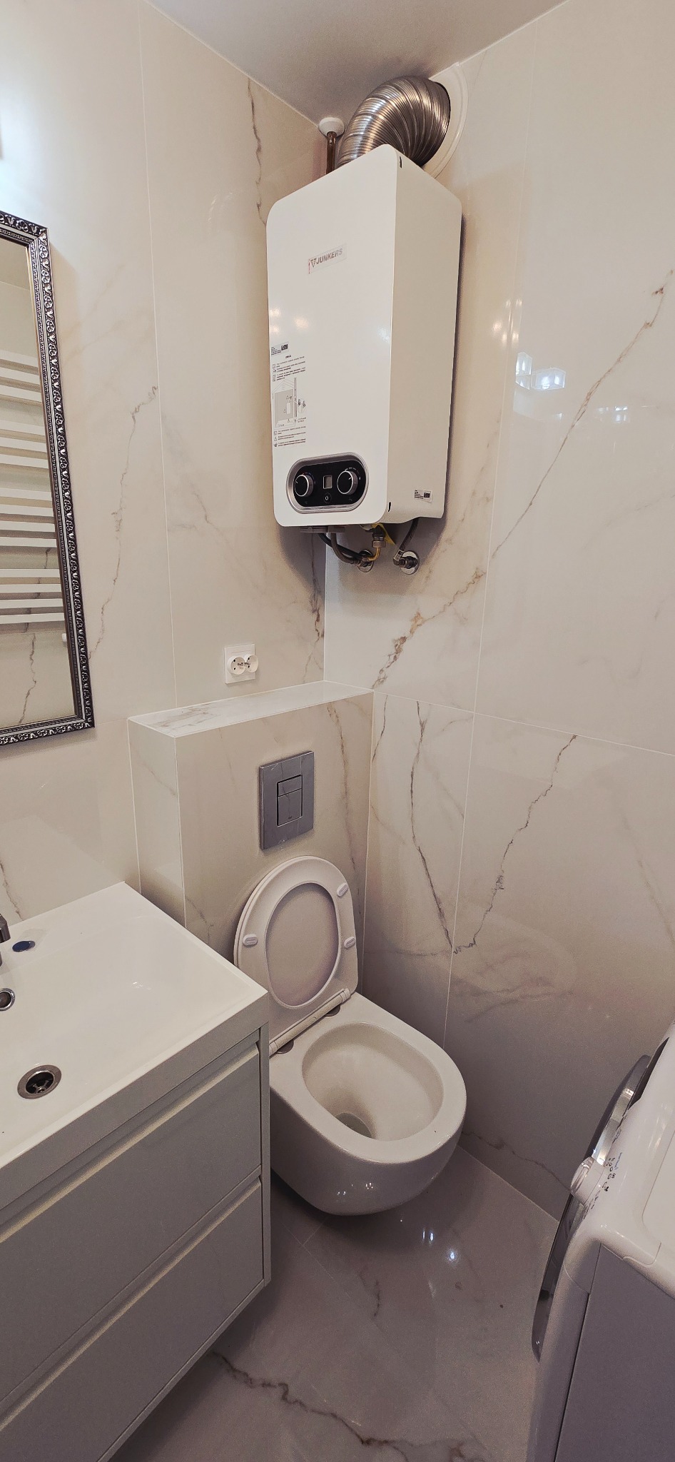 Wykończona łazienka z marmurowymi płytkami, wiszącą toaletą, szafką z umywalką i podgrzewaczem wody Junkers. Nowoczesny design.