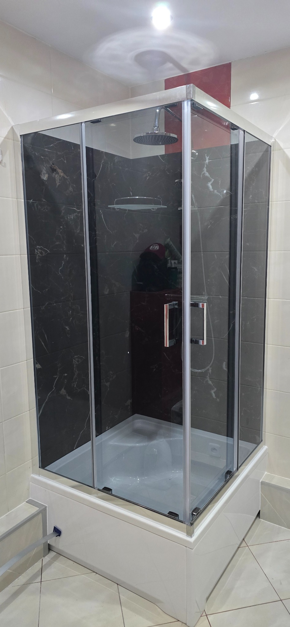 Nowoczesna kabina prysznicowa z czarnymi płytkami imitującymi marmur i deszczownicą, zamontowana na wannie. Jasne płytki na ścianach, minimalistyczny design.