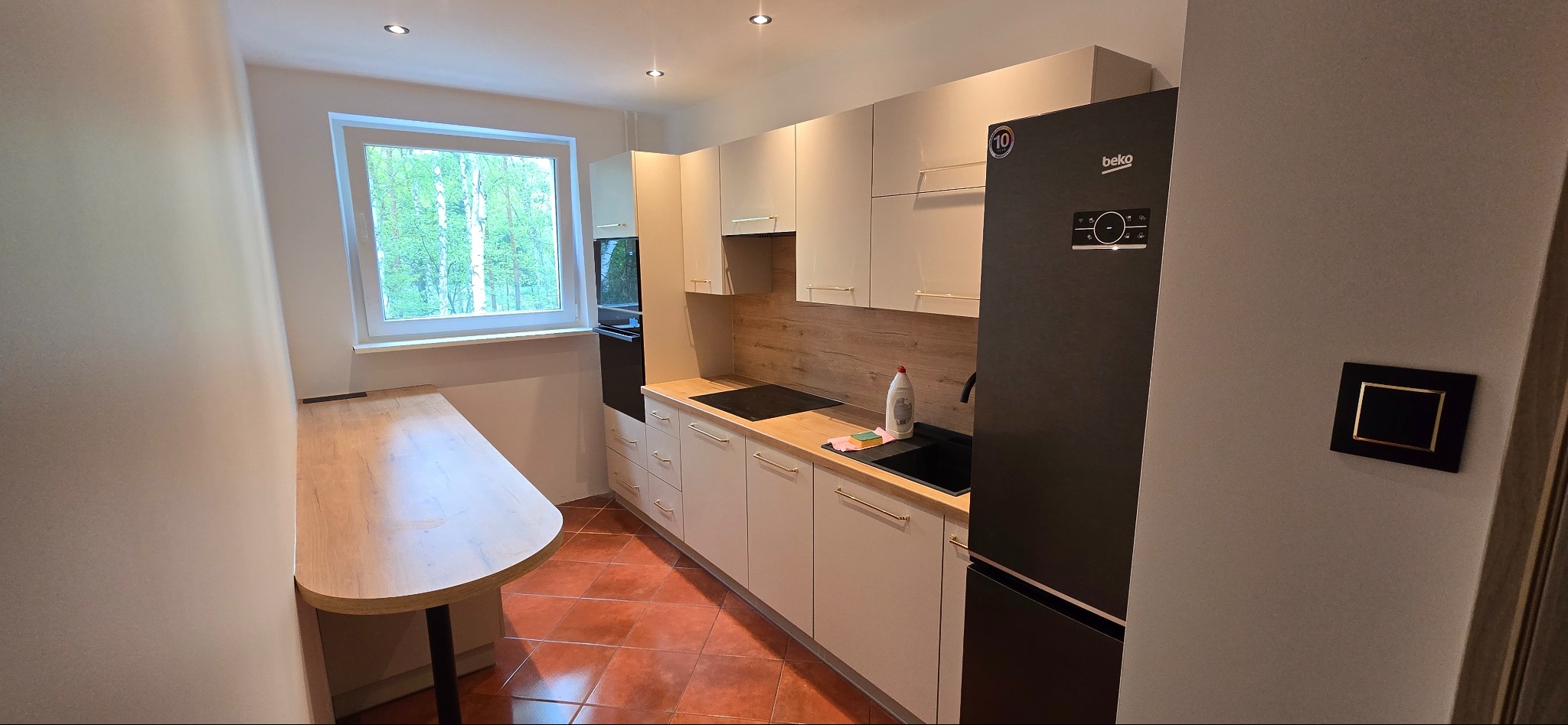 Jasna kuchnia z brązową podłogą i drewnianym blatem, widok na okno z zielenią. Nowoczesne szafki z delikatnymi uchwytami, czarna lodówka Beko.
