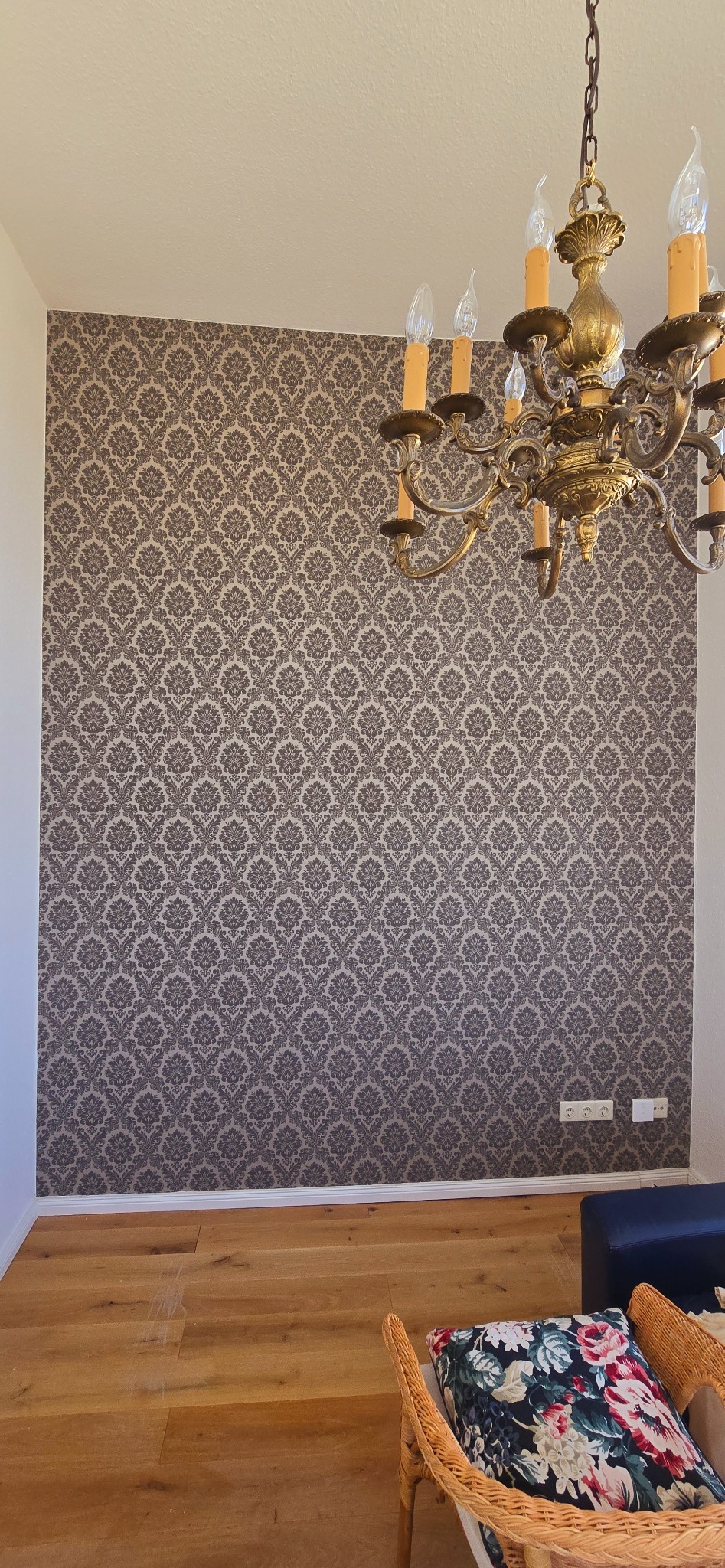 Ściana z tapetą w ornamenty w odcieniach szarości, parkiet na podłodze, fragment żyrandola i mebli. Widoczne gniazdka elektryczne przy podłodze.