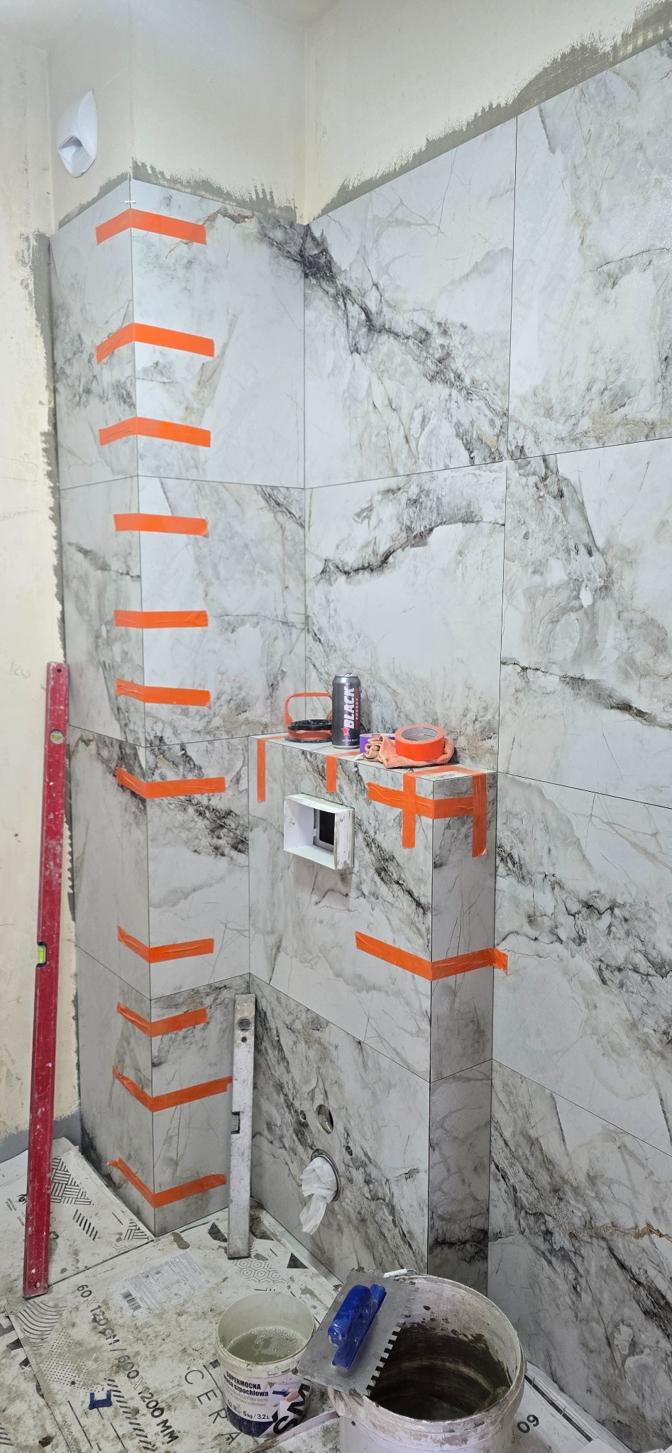 Łazienka w trakcie wykańczania: marmurowe płytki z pomarańczową taśmą, poziomice i narzędzia glazurnicze. Widoczny etap prac wykończeniowych.