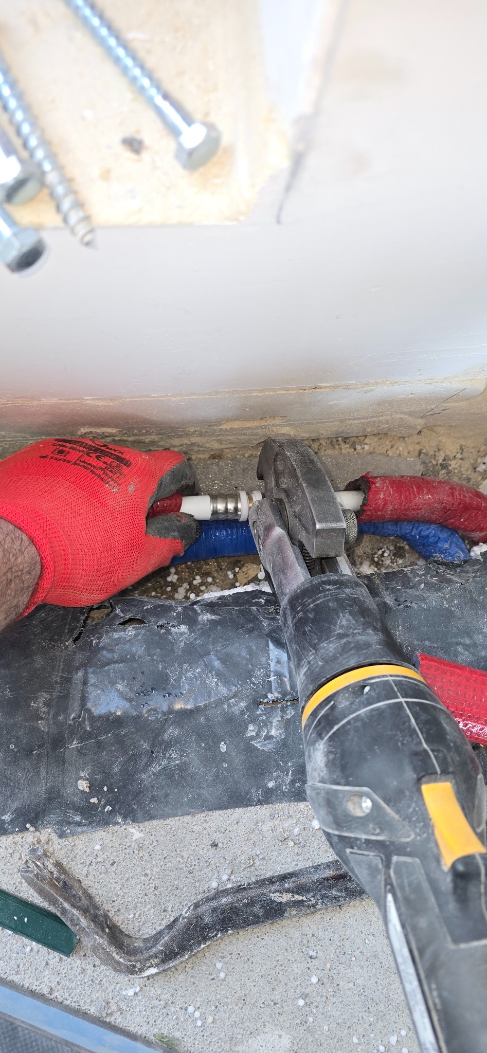 Instalacja rur PEX z użyciem zaciskarki, widoczne rury w izolacji czerwonej i niebieskiej. Praca w trakcie wykonywania, rękawice ochronne.