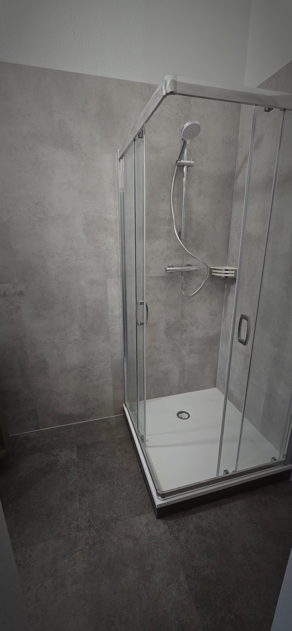 Nowoczesna kabina prysznicowa z szarymi płytkami imitującymi beton. Kabina narożna z chromowanymi detalami i ciemną podłogą. Minimalistyczny design.