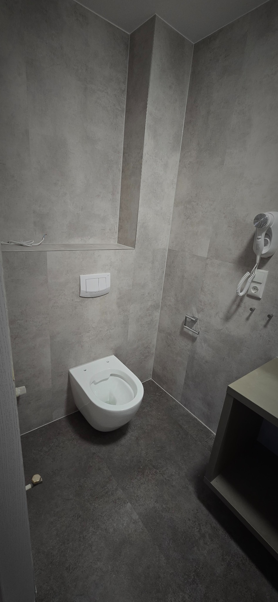 Nowoczesna łazienka w szarościach: wisząca toaleta, płytki imitujące beton, minimalistyczny design. Widoczny fragment szafki i suszarka do włosów.