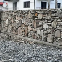 Kamienny mur ogrodzeniowy z nieregularnych, szarych i brązowych kamieni różnej wielkości, z widocznym betonowym fundamentem i gruzem na ziemi.