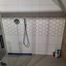 LUX-HAUS - Wykończona kabina prysznicowa z biało-czarnymi heksagonalnymi płytkami na górnej części ściany, szare płytki na dole, widoczny panel prysznicowy i słuchawka, oraz częściowo widoczna szklana ściana...