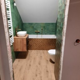 LUX-HAUS - Wnętrze łazienki na poddaszu z wanną obudowaną mozaiką, zielonymi ścianami, drewnopodobną podłogą, umywalką nablatową na drewnianej szafce i toaletą, widziane z perspektywy wejścia w trakcie remontu.