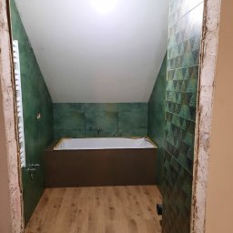 LUX-HAUS - Łazienka na poddaszu w trakcie remontu, z zielonymi płytkami na ścianach, drewnopodobną podłogą i białą wanną w zabudowie, widoczna przez surowe futryny drzwi.